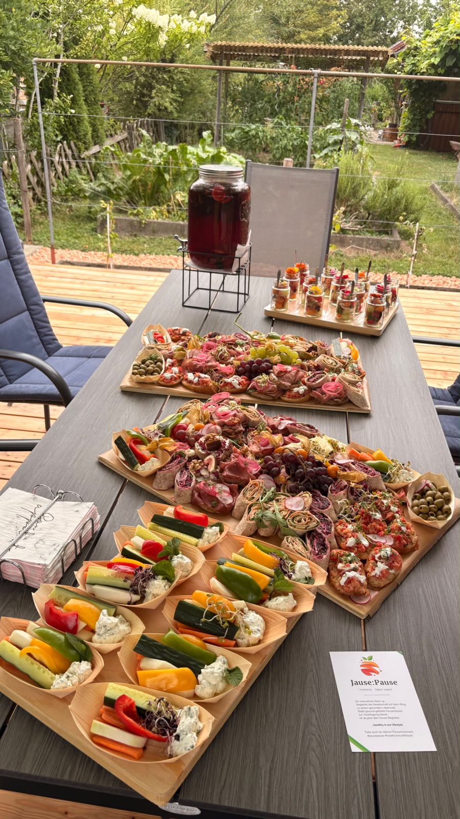 Aufgebautes Buffet von Mittags:Pause auf einem Gartentisch mit Platten, Gemüsebechern und Getränkespender